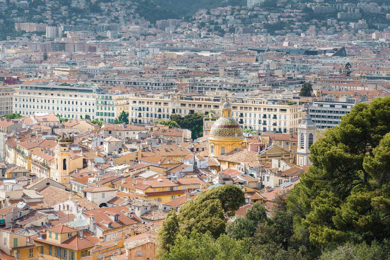 High-angle cityscape of Nice, France, showcasing the distinctive yellow-domed Cathédrale Sainte-Réparate amidst terracotta rooftops and dense urban buildings, with lush green trees in the foreground.