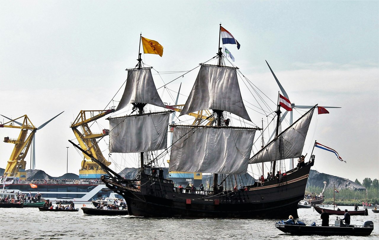 A dark-hulled replica of a historic Dutch tall ship, with white sails partially unfurled and flying Dutch and Zeeland flags, cruises a waterway. Smaller boats accompany it, with modern industrial cranes, wind turbines, and bulk cargo visible in the backgr