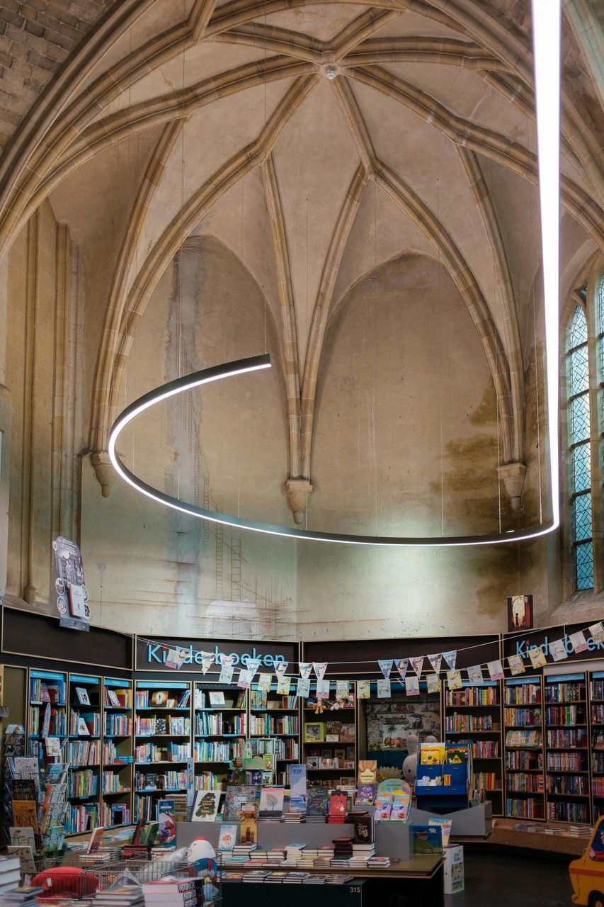 A spacious bookstore located inside a historic Gothic church in Maastricht, Netherlands, featuring high vaulted ceilings, modern circular lighting, and shelves filled with a wide variety of books, including a visible "Kinderboeken" (children's books) sect