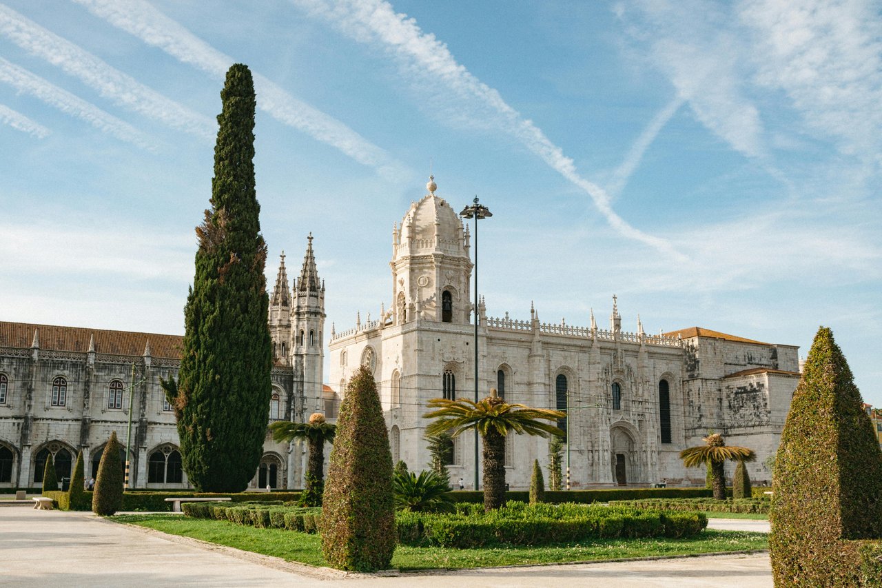 Historic Jerónimos Monastery in Belém, Lisbon, Portugal, featuring grand Manueline architecture with intricate white stone details, multiple spires, and a prominent central dome, set against a blue sky with streaky clouds. The foreground includes manicure