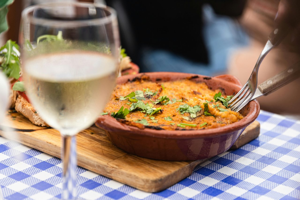 Close-up of a rustic baked casserole or gratin dish with fresh herbs, accompanied by a glass of white wine and toasted bread, on a blue and white gingham tablecloth. A hand scoops food from the terracotta bowl, suggesting a delightful Mediterranean dining