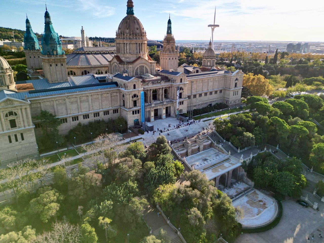 Aerial view of the National Art Museum of Catalonia (MNAC) on Montjuïc hill in Barcelona, Spain, featuring its majestic Neo-Baroque architecture with domes and towers, grand staircases, and surrounding lush parkland under a sunny sky, with the city skylin