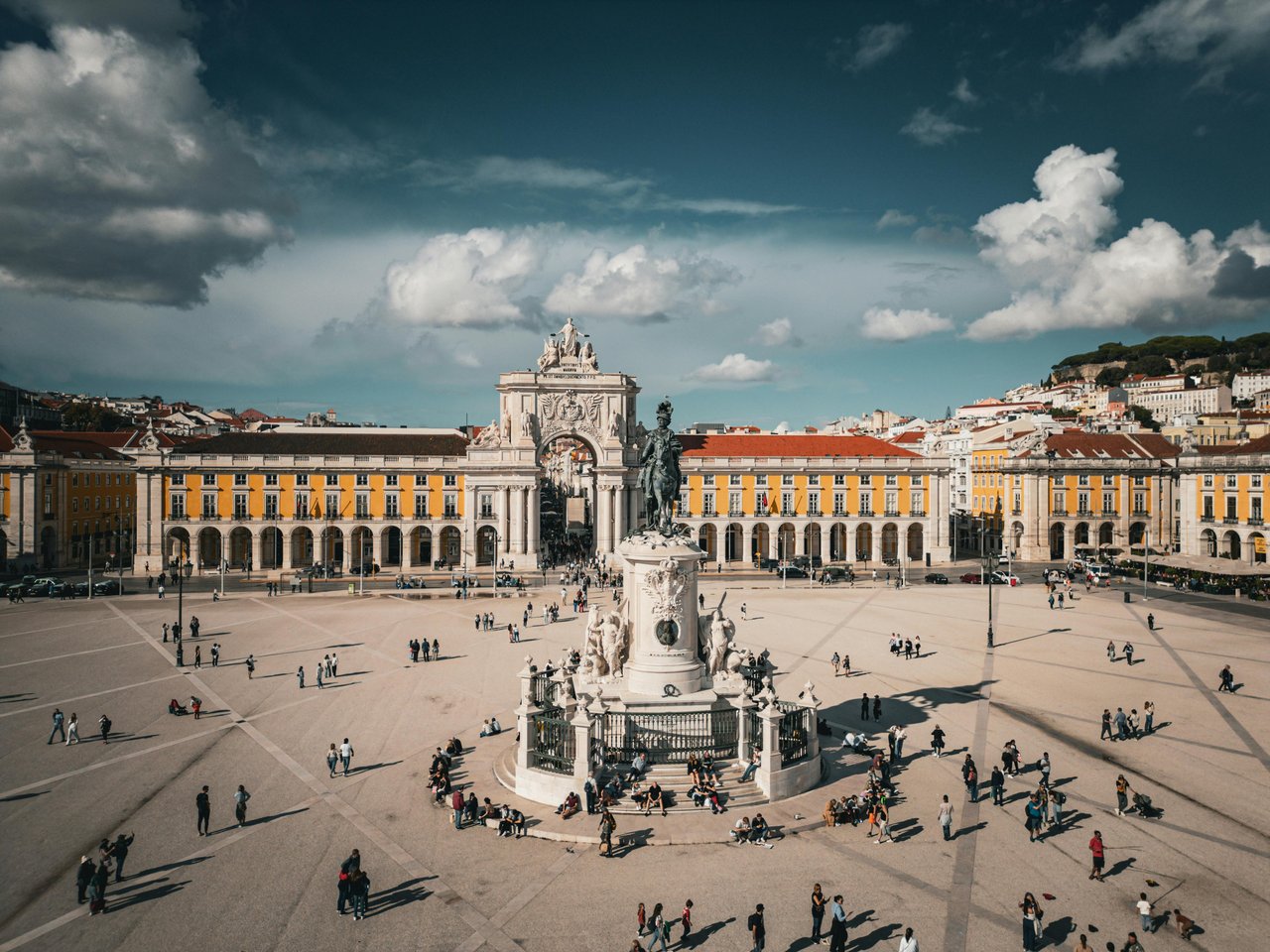 Lisbon, Portugal: Praça do Comércio square with the equestrian statue of King José I, the grand Rua Augusta Arch, and historic yellow buildings bustling with people under a dramatic sky.