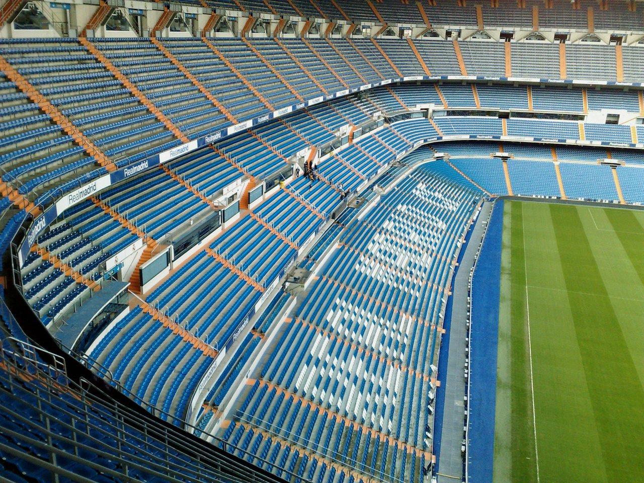 High-angle view of the iconic Santiago Bernabéu Stadium in Madrid, Spain, home of Real Madrid. The stadium features rows of blue and orange seats, with a prominent section of white seats spelling out 'REAL MADRID' on the lower tier. A lush green football 