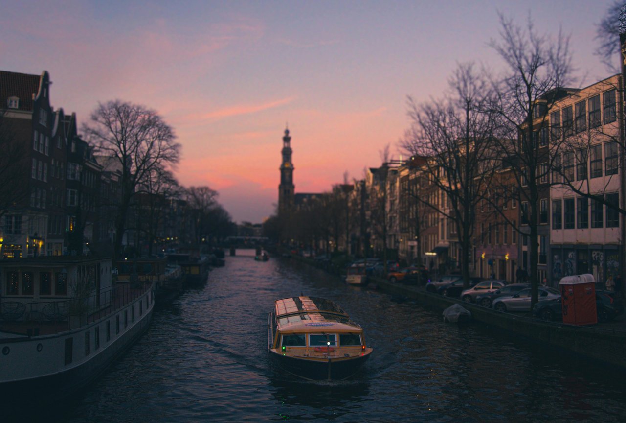 Scenic Amsterdam canal at sunset, featuring a tour boat navigating the waterway flanked by historic Dutch canal houses and the iconic Westerkerk tower under a vibrant purple and pink sky.
