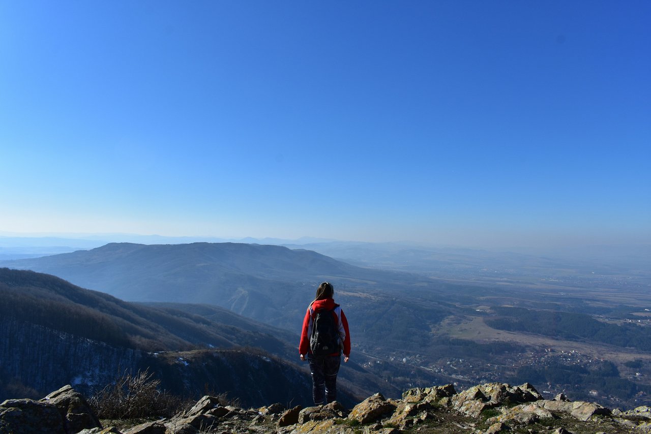 A hiker stands on a rocky peak in the Balkan Mountains, Bulgaria, looking out over a sprawling panoramic view of layered mountains and a valley with a town under a bright blue sky.