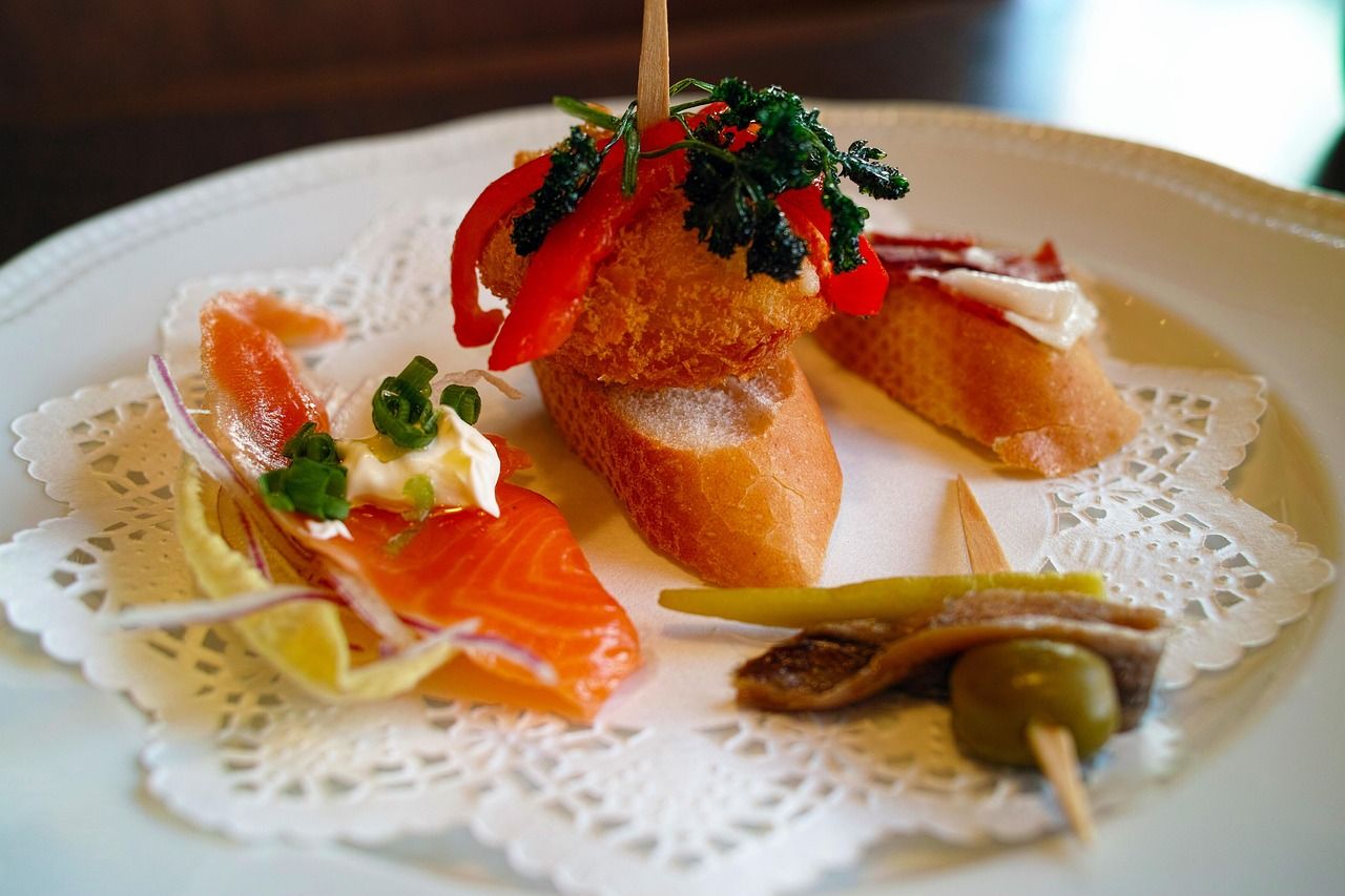 A selection of gourmet Spanish pintxos on a white plate with a doily, featuring smoked salmon, a fried croquette with red peppers, and an anchovy with olive gilda.