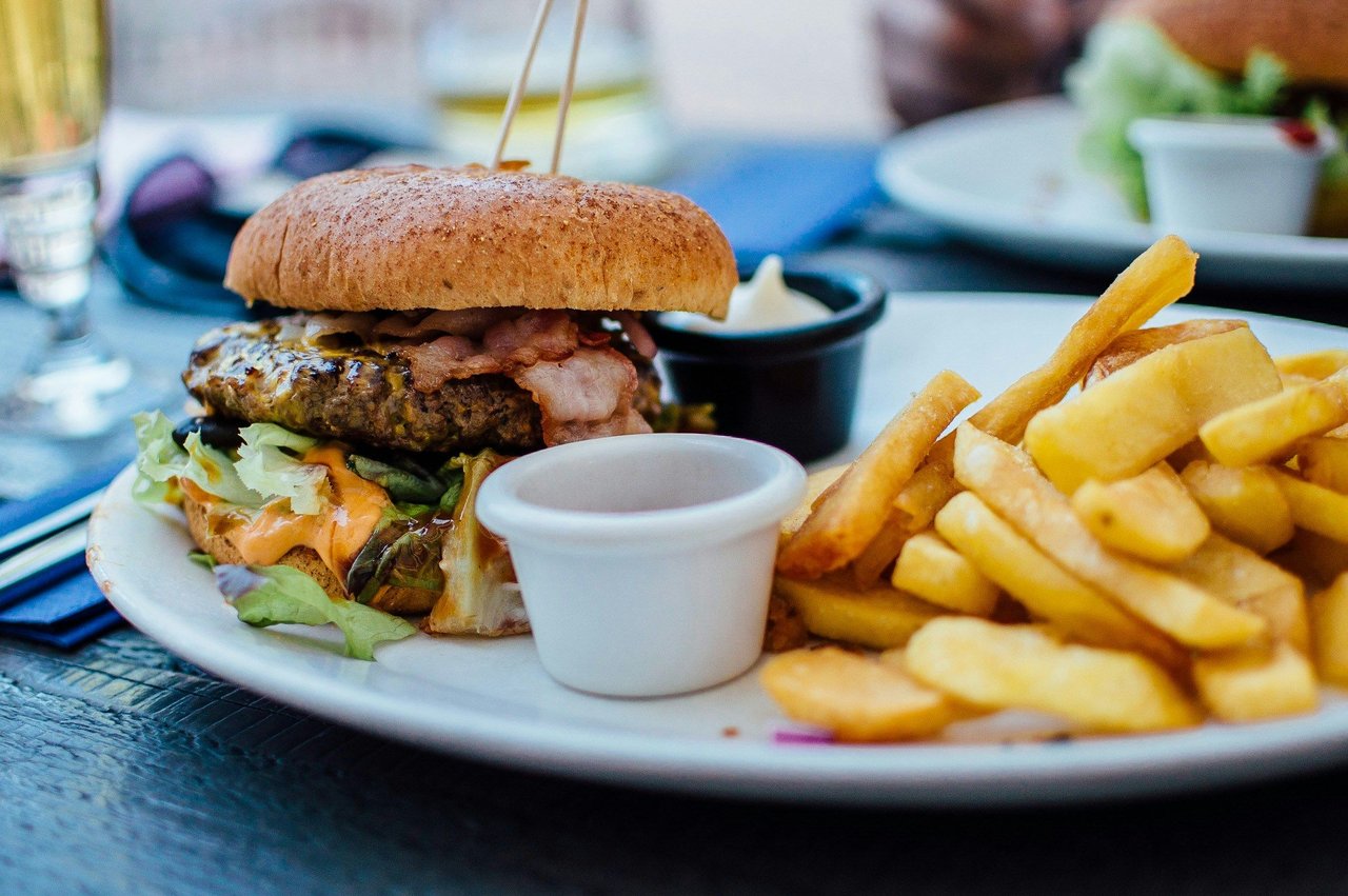 Gourmet bacon burger with lettuce and a creamy orange sauce, served with golden french fries and two dipping sauce ramekins on a white plate at a restaurant table.