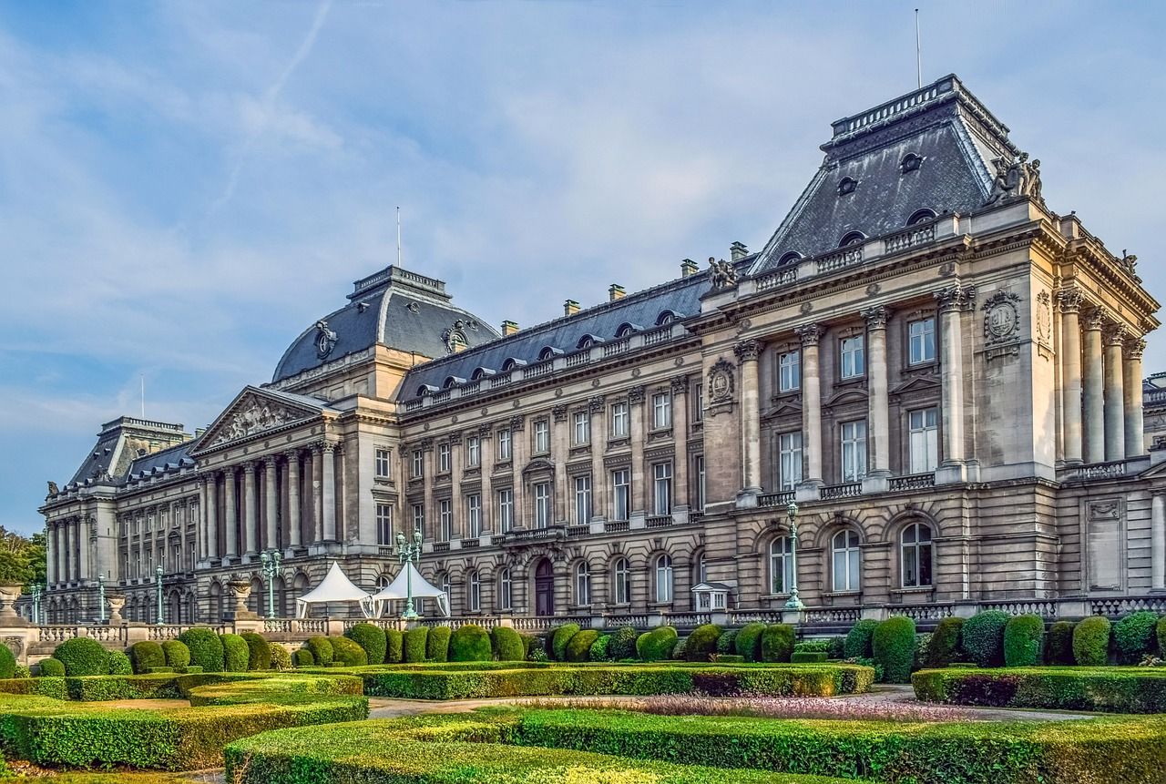 The majestic Royal Palace of Brussels in Belgium, showcasing its classical architecture with tall columns and intricate details, set against a blue sky with manicured green formal gardens in the foreground.