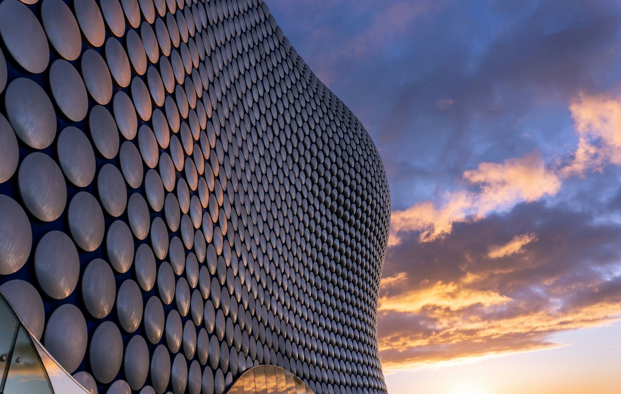 The iconic Selfridges building in Birmingham, UK, featuring its curved facade covered in thousands of silvery discs, set against a vibrant blue and orange sunset sky.