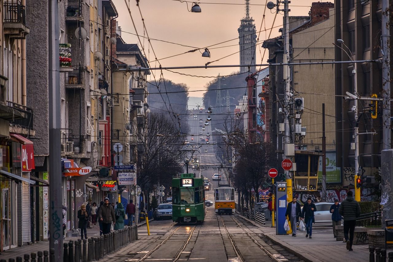 Bustling street scene in Sofia, Bulgaria, with two trams on tracks, historic buildings, pedestrians, and the Kopitoto TV Tower visible in the distance.