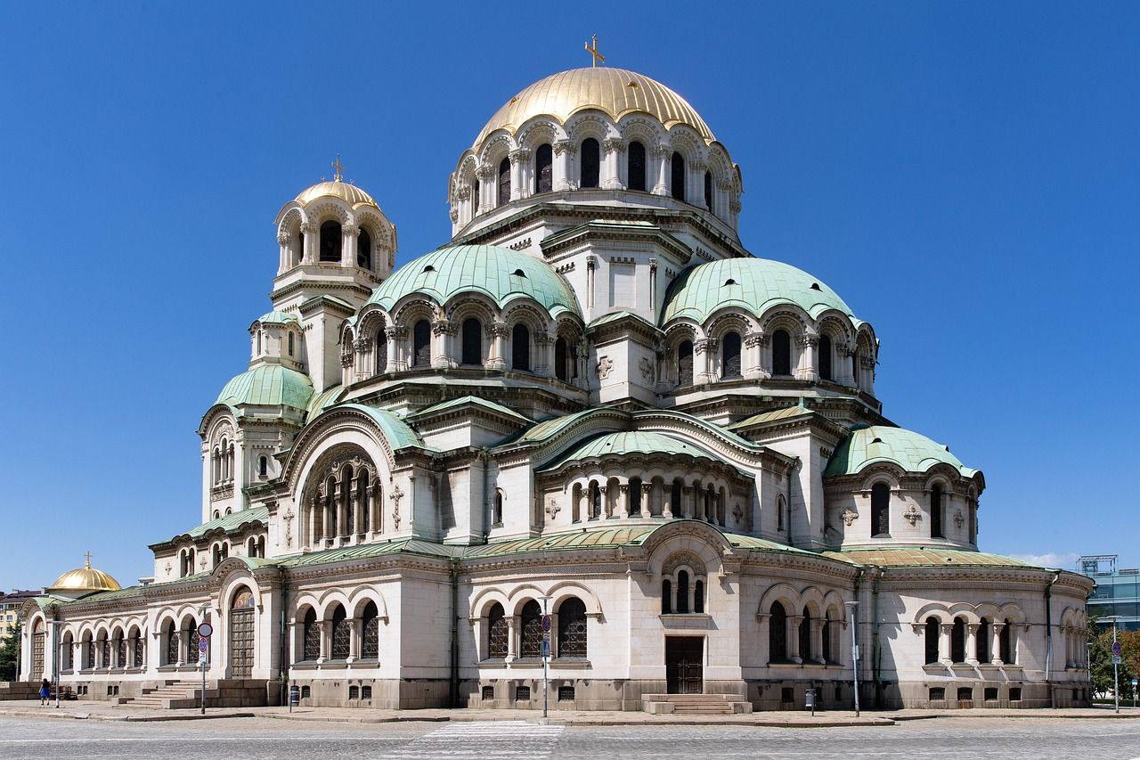 Alexander Nevsky Cathedral in Sofia, Bulgaria, with its grand white facade, golden central dome, and multiple green domes against a clear blue sky.