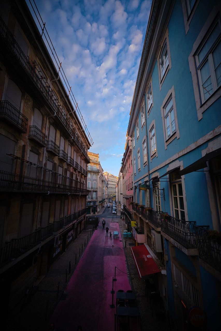 A vibrant scene of Lisbon's iconic Pink Street (Rua Nova do Carvalho), featuring its distinctive magenta pavement flanked by a bright blue building and traditional facades under a partly cloudy sky.