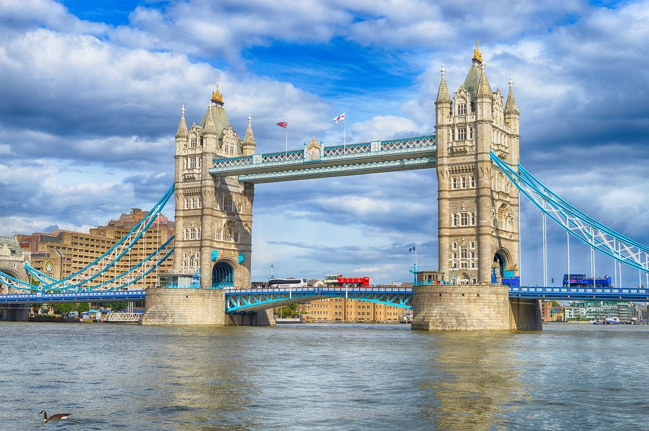 Tower Bridge in London, a historic landmark with grey stone towers and blue suspension cables, spanning the River Thames. Red double-decker buses cross the bridge under a partly cloudy sky, with a duck swimming in the foreground.