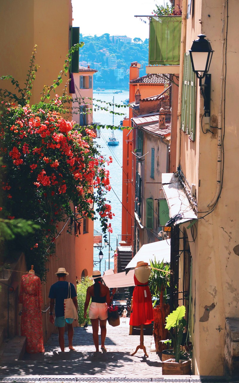 A sun-drenched narrow cobblestone street in a Mediterranean coastal town, with a couple walking towards a serene turquoise bay. Vibrant red bougainvillea cascades from colorful buildings with green shutters, and boats are visible on the water below.