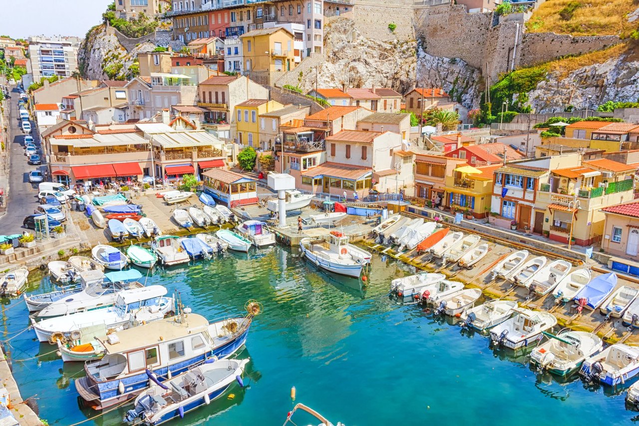Scenic view of Vallon des Auffes fishing port in Marseille, France, showcasing colorful Mediterranean houses, clear turquoise water, and numerous small boats docked along the picturesque rocky cove.