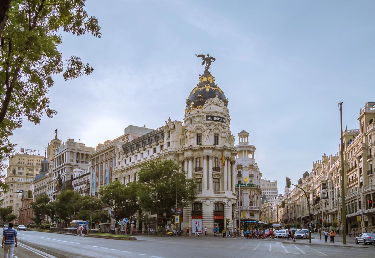 The iconic Metropolis Building with its distinctive dome and statue at the intersection of Gran Vía and Calle de Alcalá in Madrid, Spain, bustling with pedestrians and cars on a clear day.