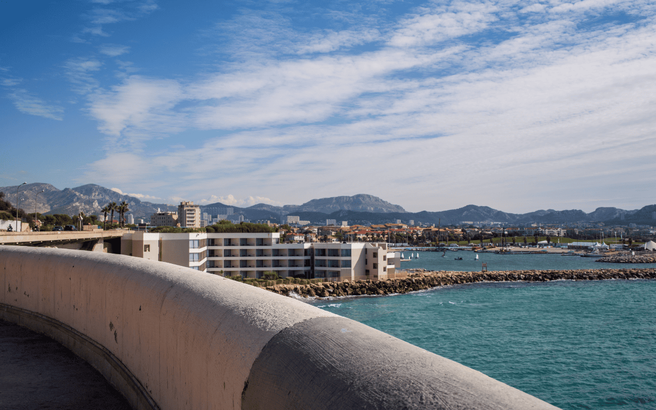 Panoramic view of Marseille, France, featuring turquoise Mediterranean waters, a marina with boats, modern coastal buildings, and distant rugged limestone mountains under a blue sky.