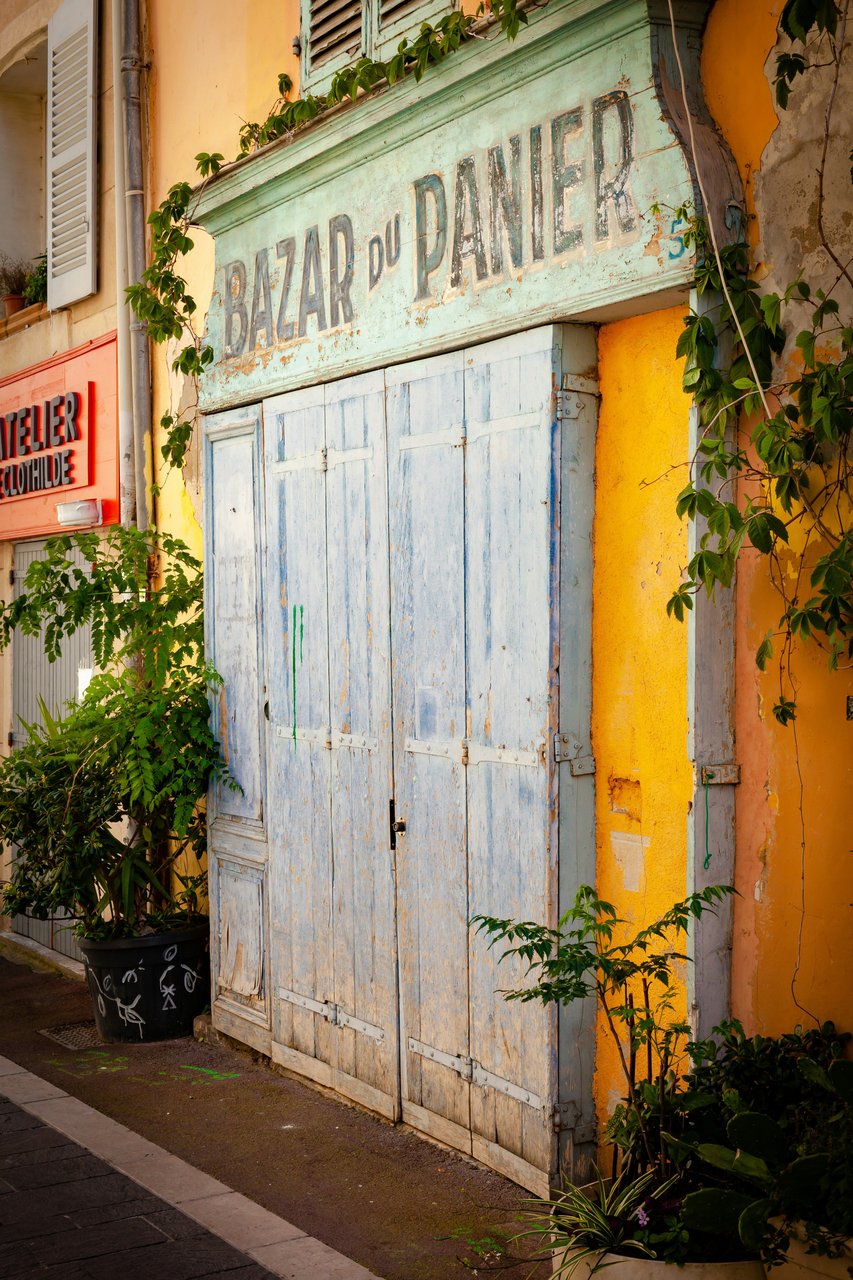 Colorful old shop facade in Le Panier, Marseille, France, featuring weathered blue wooden doors, a faded "Bazar du Panier" sign, vibrant yellow and orange walls, and climbing green vines with potted plants.