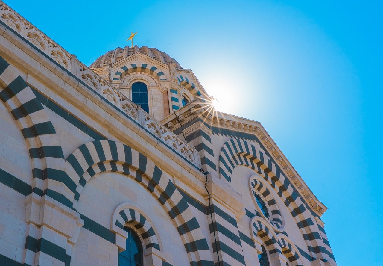 Notre-Dame de la Garde Basilica, Marseille, France, featuring its distinctive striped stone architecture, dome, and golden cross under a bright blue sky with sun rays.