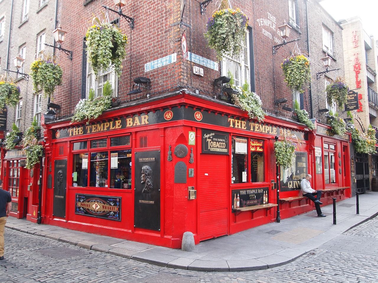The iconic bright red Temple Bar pub, established 1840, on a cobblestone corner street in Dublin, Ireland, adorned with lush hanging baskets.
