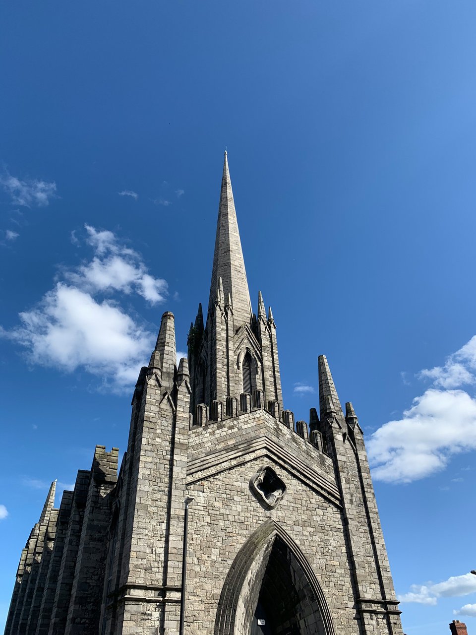 Low-angle view of the historic St. Patrick's Church in Belfast, Northern Ireland, showcasing its tall stone spire and Gothic Revival architecture against a bright blue sky with scattered white clouds.