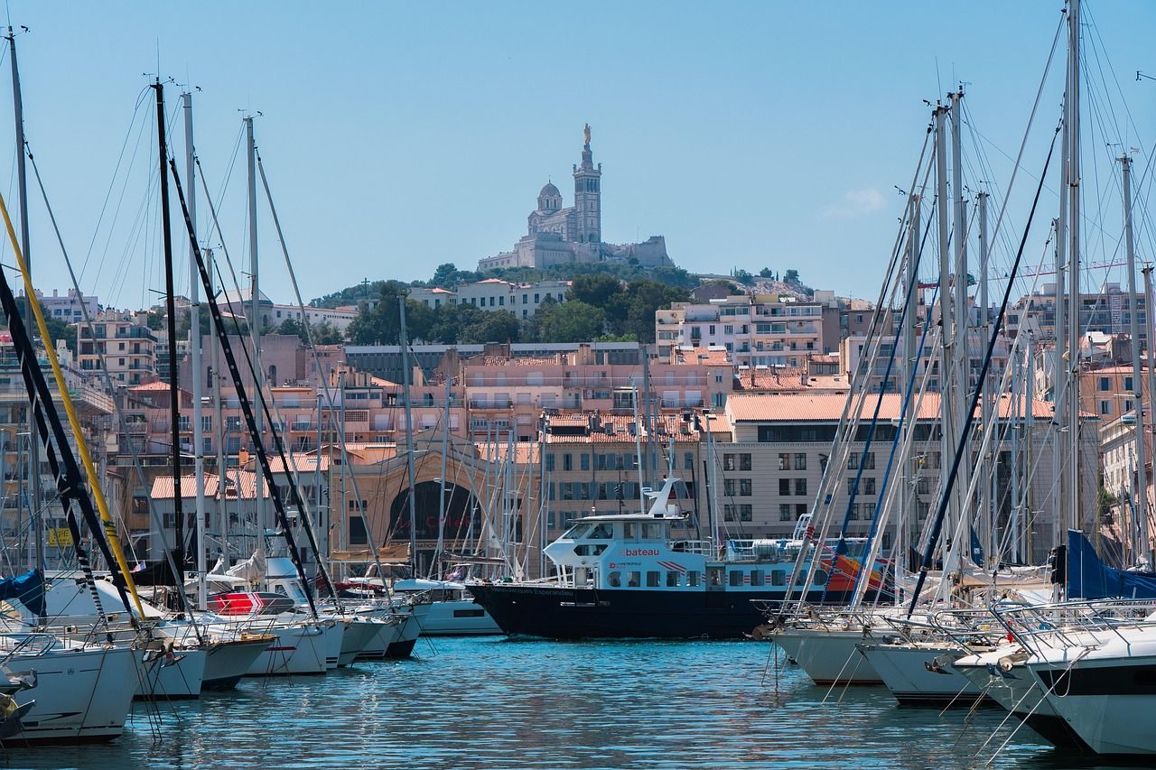 A vibrant scene of Marseille's Vieux Port (Old Port) with numerous sailboats and a tour boat, framed by city buildings, under a clear blue sky, with the iconic Notre-Dame de la Garde basilica prominently on a hill in the background.