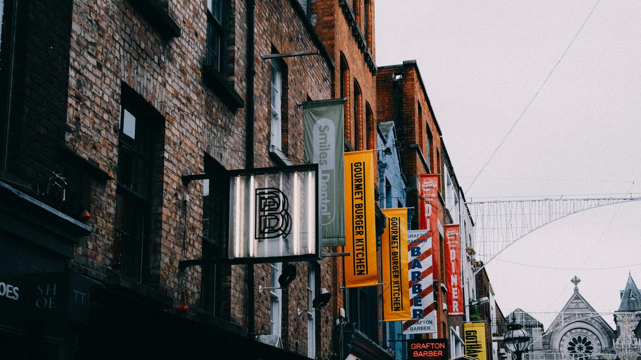 A bustling street scene on Grafton Street, Dublin, Ireland, featuring historic red brick buildings with prominent hanging signs for businesses including 'Gourmet Burger Kitchen' and 'Grafton Barber' under an overcast sky.