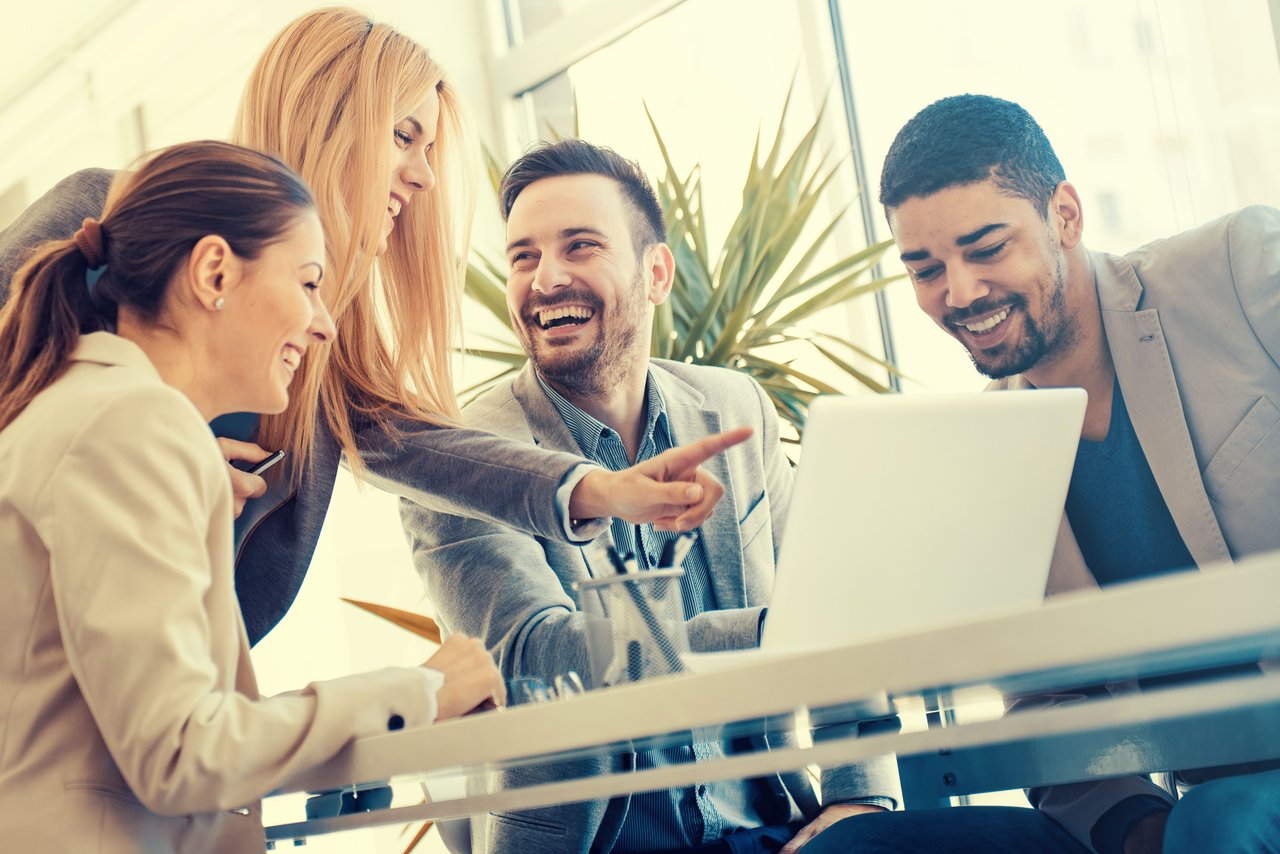 Diverse group of four happy business professionals collaborating and laughing around a laptop during a team meeting in a bright modern office.