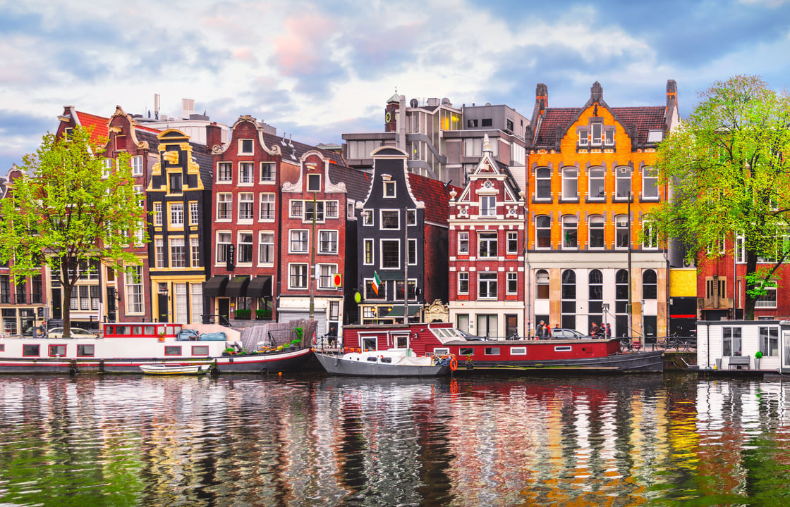 Colorful traditional Dutch canal houses lining a tranquil Amsterdam canal with houseboats and reflections on the water under a cloudy sky.