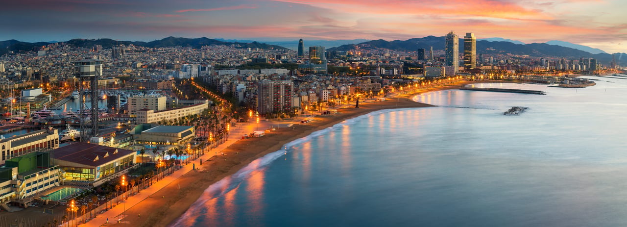 Panoramic night view of Barcelona, Spain, showcasing the illuminated coastline, city skyline, and urban beach with the Mediterranean Sea at dusk.