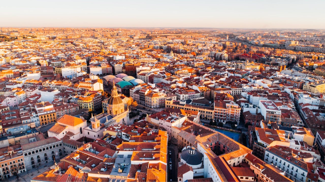 Aerial panoramic view of Madrid, Spain, at golden hour, featuring historic buildings, red-tiled rooftops, and iconic landmarks like Mercado de San Antón.