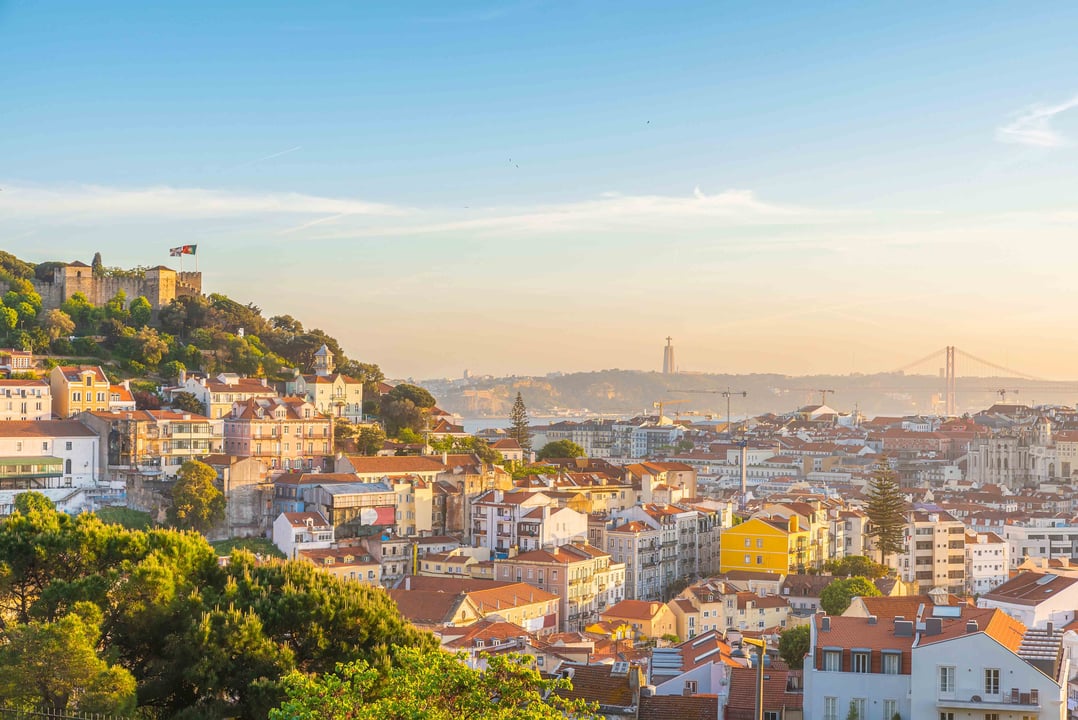 Cityscape with red-roofed buildings, hilltop castle, and distant suspension bridge.