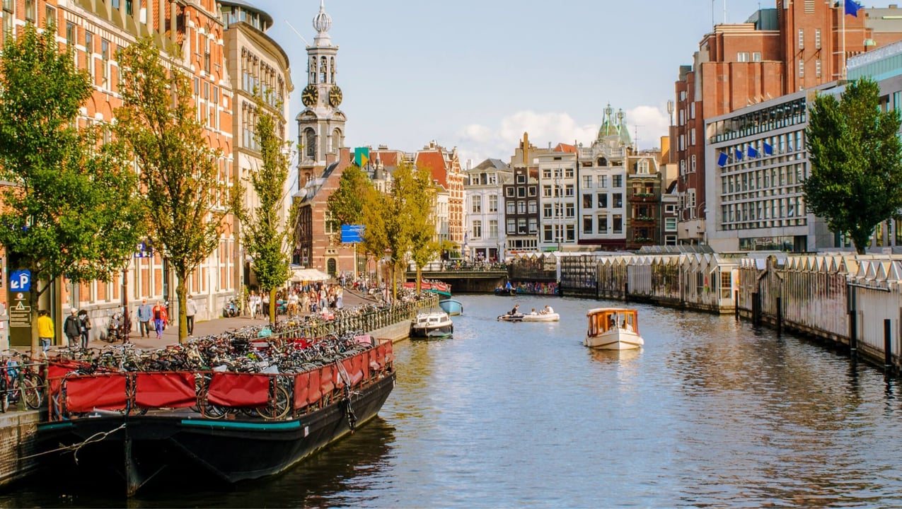 Vibrant cityscape of Amsterdam with a canal, historic gabled buildings, and numerous bicycles parked on houseboats. Boats cruise the water, and the iconic Zuiderkerk tower rises in the background.