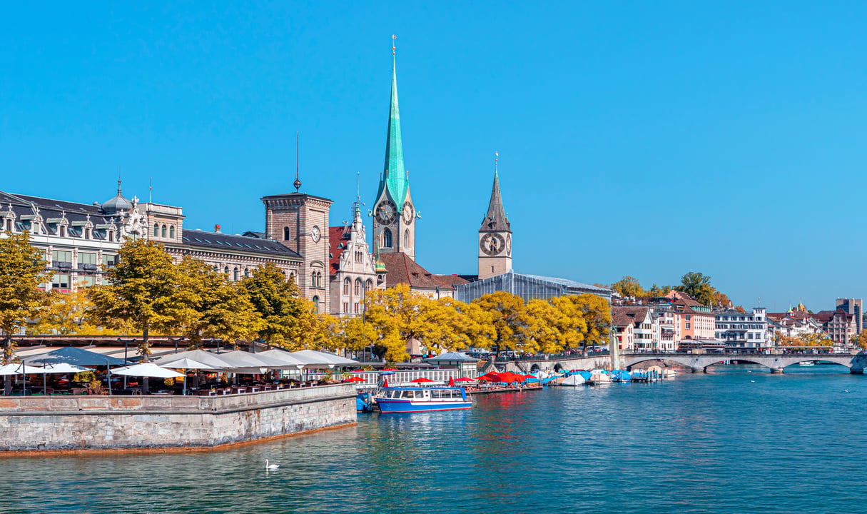 Zurich Old Town cityscape in autumn, featuring the Limmat River, historic buildings, Fraumünster Church with its green spire, and St. Peter's Church with its large clock face, under a clear blue sky.