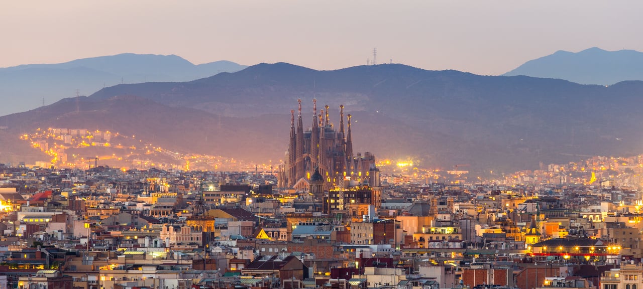 Panoramic view of the Sagrada Familia basilica towering over the illuminated Barcelona cityscape at dusk, with distant mountains.