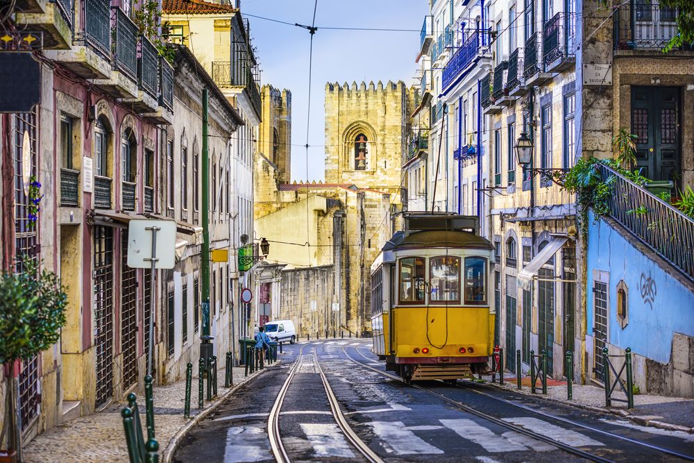 Vibrant street scene in Lisbon, Portugal, featuring a classic yellow tram, colorful historic buildings, and the Sé de Lisboa cathedral in the background.