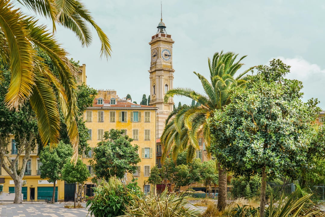 Clock tower of the Palais de la Préfecture in Nice, France, framed by lush palm trees and historic yellow buildings.