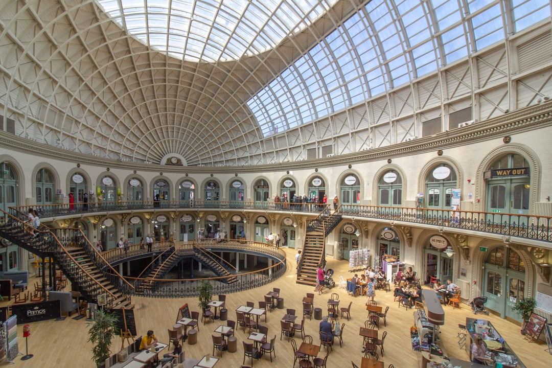 Interior of the grand, circular Leeds Corn Exchange, showcasing its impressive glass-domed roof, ornate Victorian architecture with multiple levels of arched shopfronts, and elegant staircases. Visitors are visible enjoying cafes and browsing boutiques wi