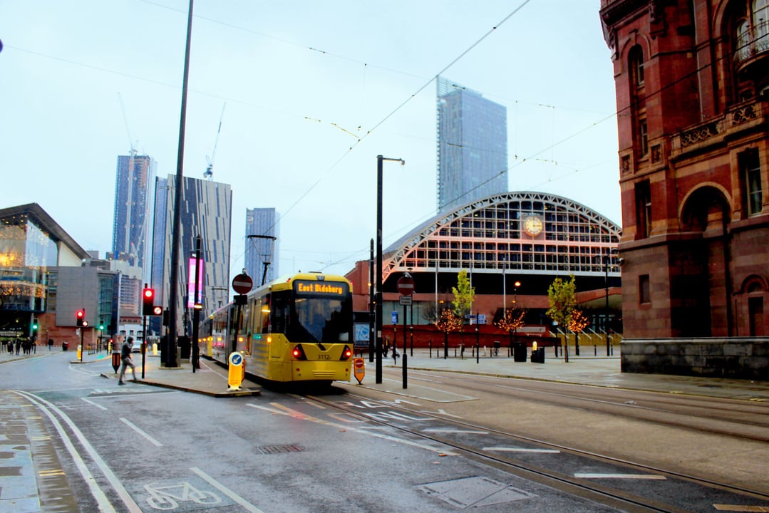Manchester city street scene with a yellow Metrolink tram heading towards East Didsbury, passing Manchester Central Convention Complex and modern skyscrapers on a wet day.