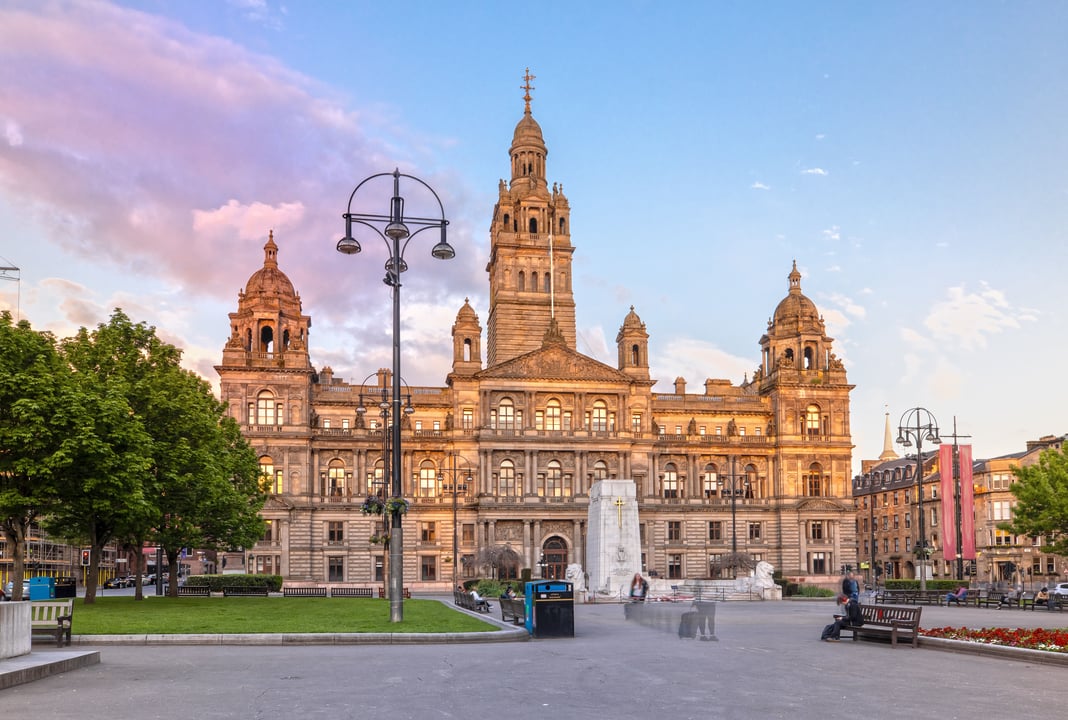 Ornate Victorian architecture of Glasgow City Chambers and George Square in Scotland at sunset, featuring the grand building, war memorial, and public park under a colorful sky.