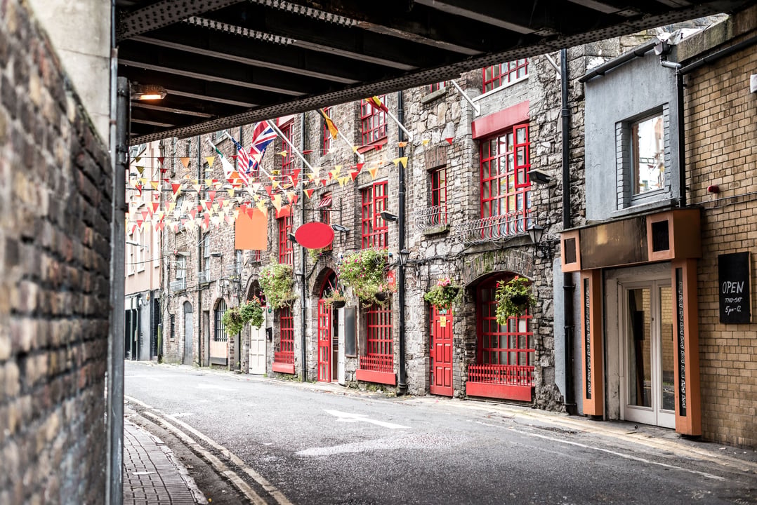 Historic alleyway in Dublin's Temple Bar district, featuring charming stone buildings with bright red windows and doors, adorned with colorful bunting, viewed from under a dark archway.