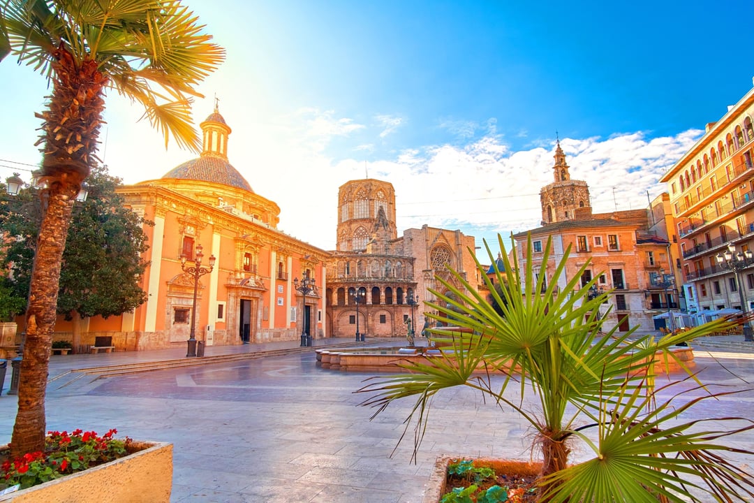 Vibrant Plaza de la Virgen in Valencia, Spain, with the Miguelete bell tower, Valencia Cathedral, and Basilica de la Mare de Déu dels Desemparats under a bright blue sky, featuring palm trees and a fountain.
