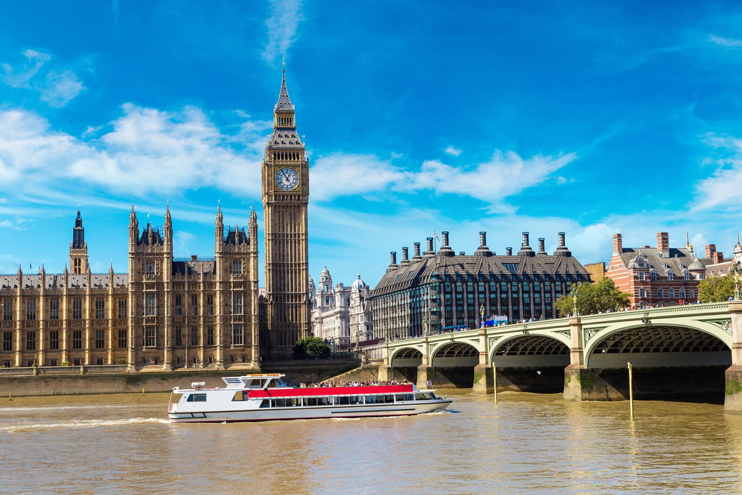 Historic Big Ben and Houses of Parliament in London, UK, beside the River Thames with a riverboat and Westminster Bridge on a clear summer day.