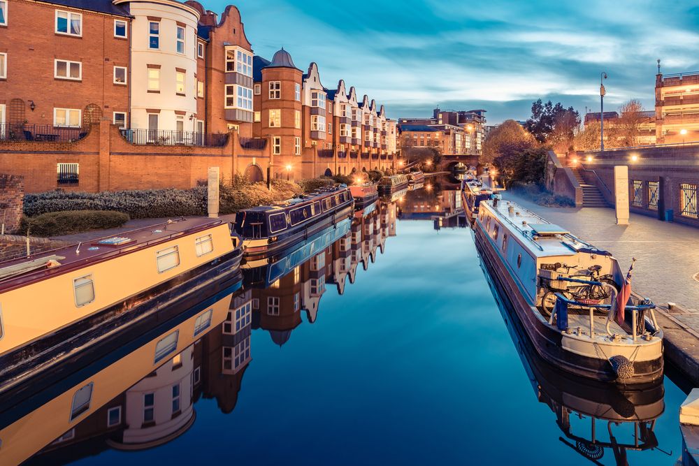 Birmingham canal network at dusk with illuminated historic brick buildings and traditional narrowboats moored along the reflective water.