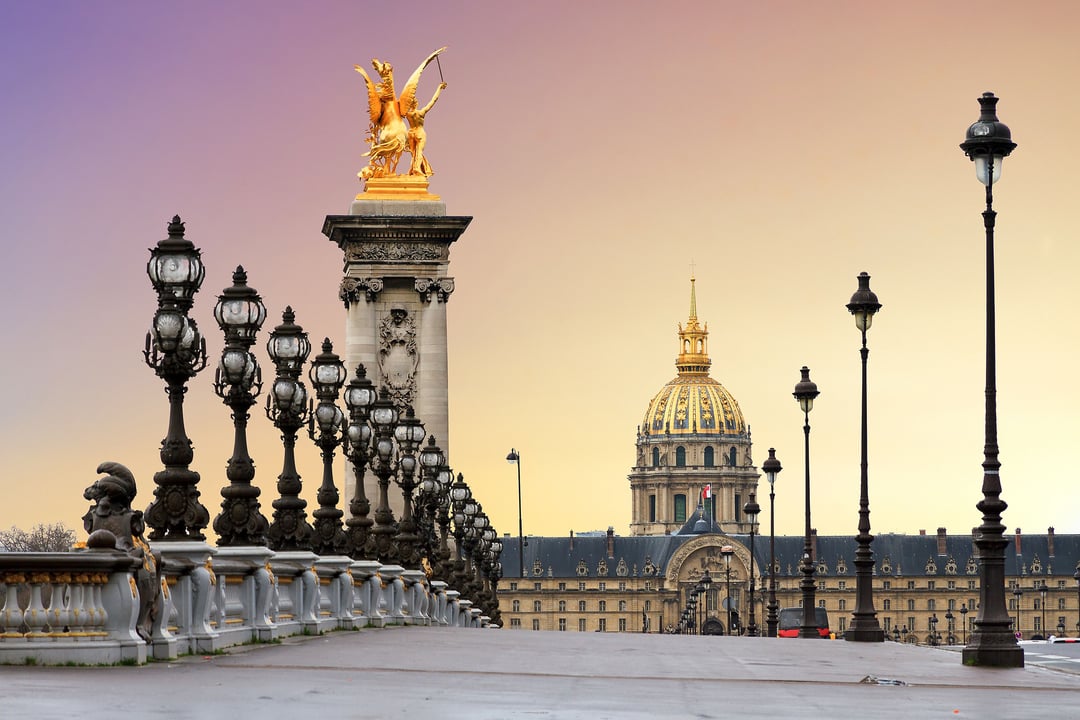 Pont Alexandre III bridge in Paris with a golden statue and ornate lampposts, facing the gilded dome of Les Invalides at sunset.