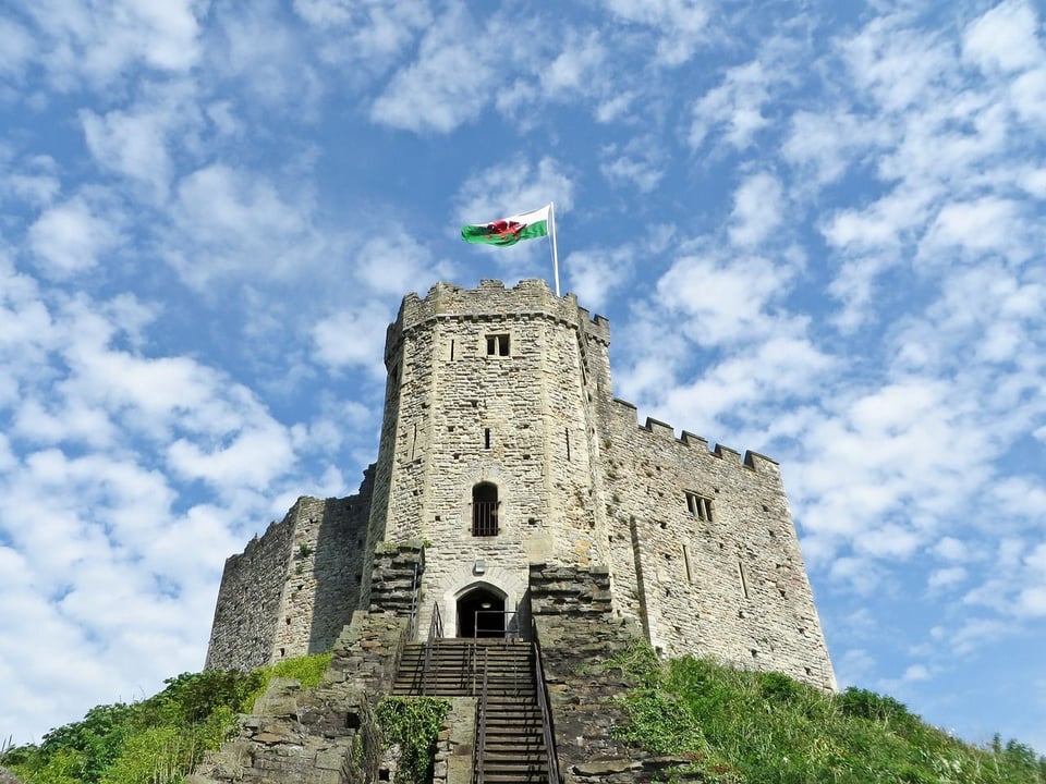 Cardiff Castle's historic Norman stone keep with the Welsh flag flying against a bright blue, cloudy sky. Stone steps ascend the grassy mound to the castle entrance.