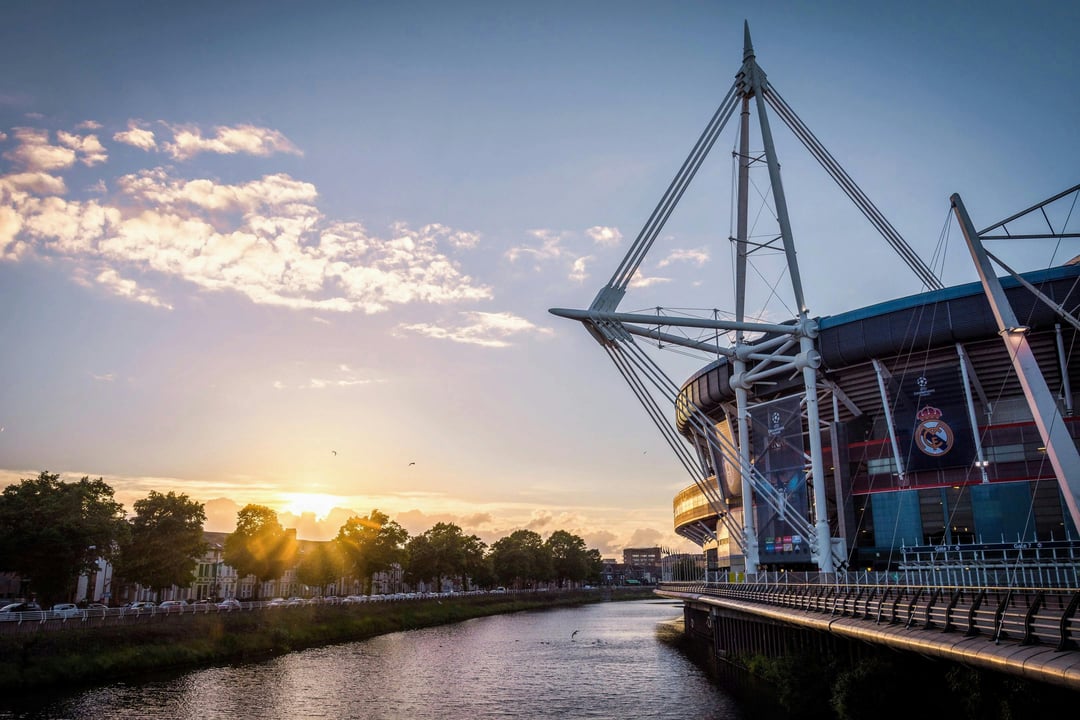 Principality Stadium in Cardiff, Wales, also known as Millennium Stadium, at sunset. The iconic sports venue, adorned with Real Madrid and Champions League banners, stands beside the River Taff under a vibrant sky with golden light and clouds.