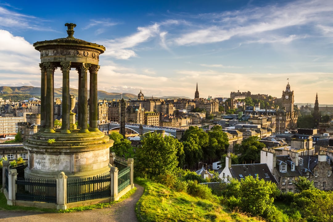 Panoramic view of Edinburgh, Scotland, featuring the classical Dugald Stewart Monument on Calton Hill, with the historic cityscape and Edinburgh Castle in the background under a golden sky.