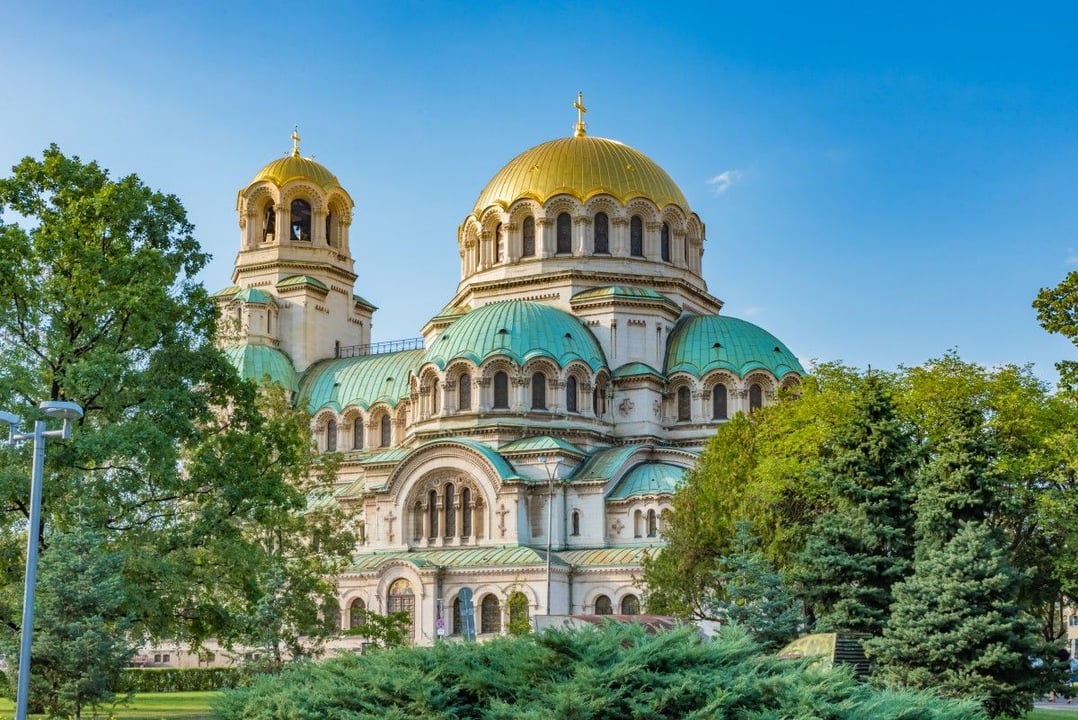 Alexander Nevsky Cathedral in Sofia, Bulgaria, featuring its iconic golden and green domes amidst lush greenery under a clear blue sky.